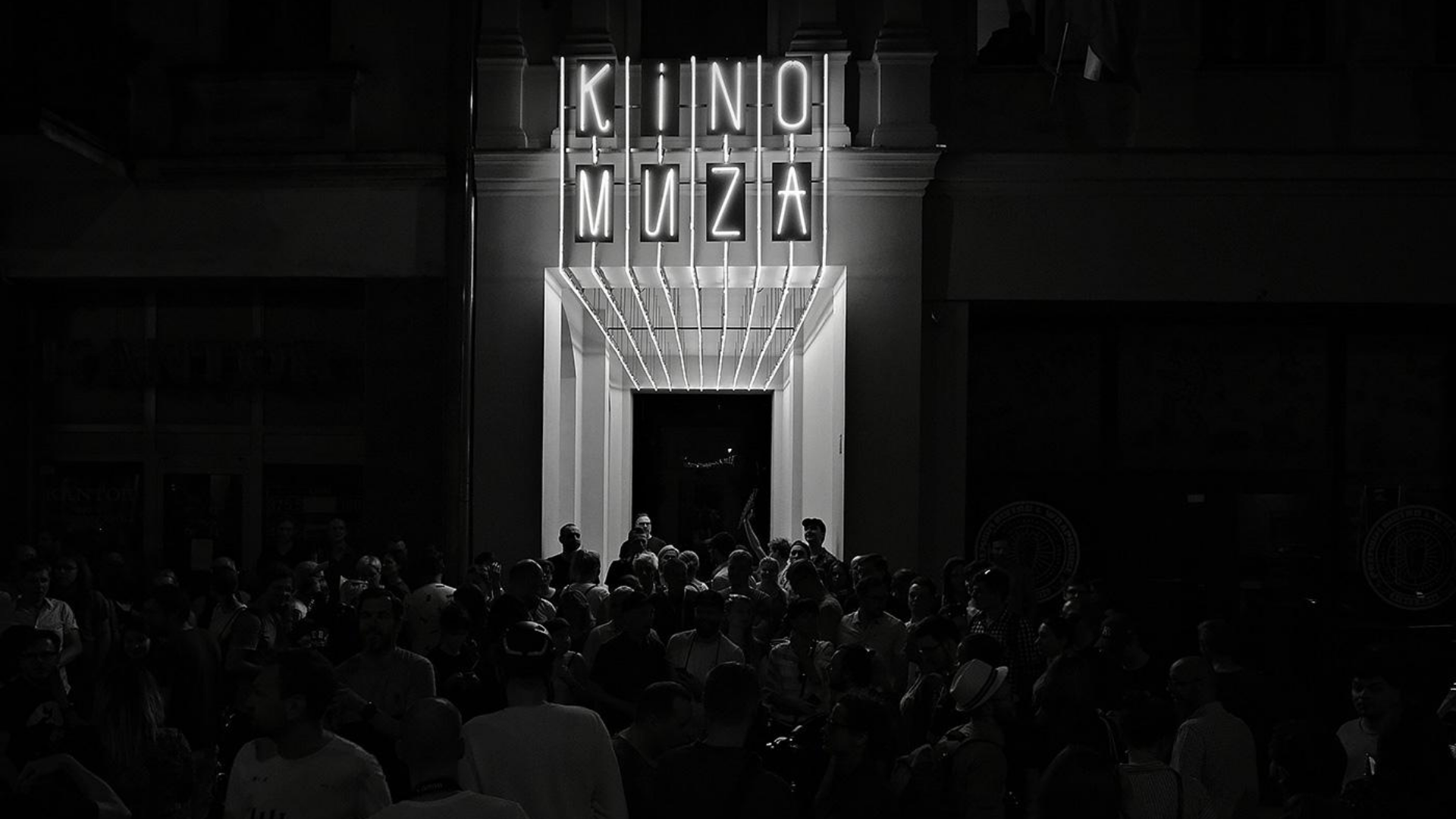 Ciemna fotofrafia przedstawia tłum przy wejściu do kina, nad wejściem świecący neon KINO MUZA.