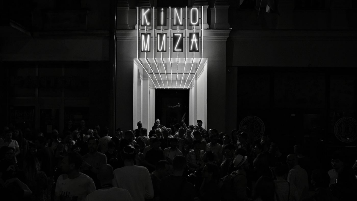 Ciemna fotofrafia przedstawia tłum przy wejściu do kina, nad wejściem świecący neon KINO MUZA.