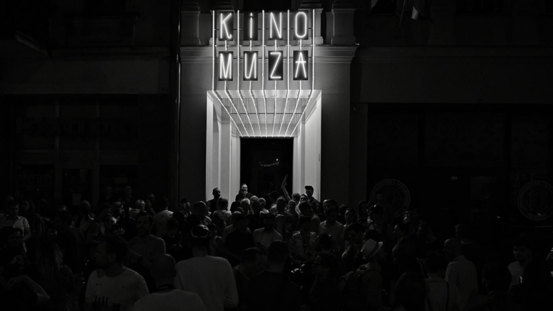 Ciemna fotofrafia przedstawia tłum przy wejściu do kina, nad wejściem świecący neon KINO MUZA.