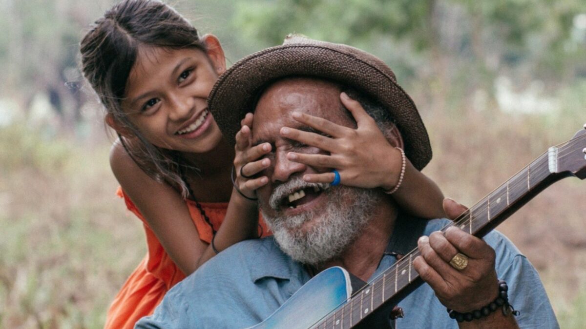 Starszy mężczyzna grający na gitarze i mała dziewczynka zasłaniająca mu oczy.