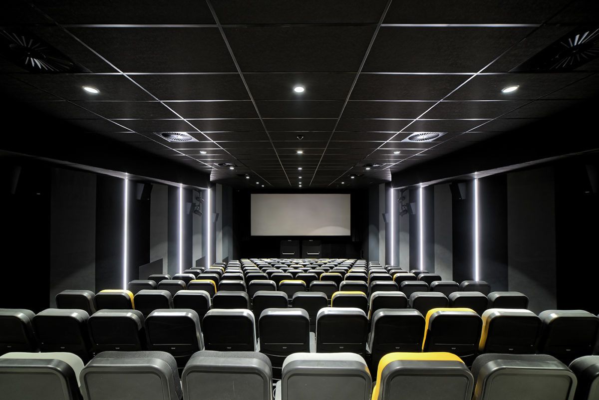 Duża stopniowana sala kinowa. Na pierwszym planie widoczne są rzędy krzeseł, w głębi widać duży biały ekran. Krzesła są w różnych kolorach, ułożonych w nieregularny wzór. Sala jest oświetlona punktowymi światłami, osadzonymi w ciemnym suficie.