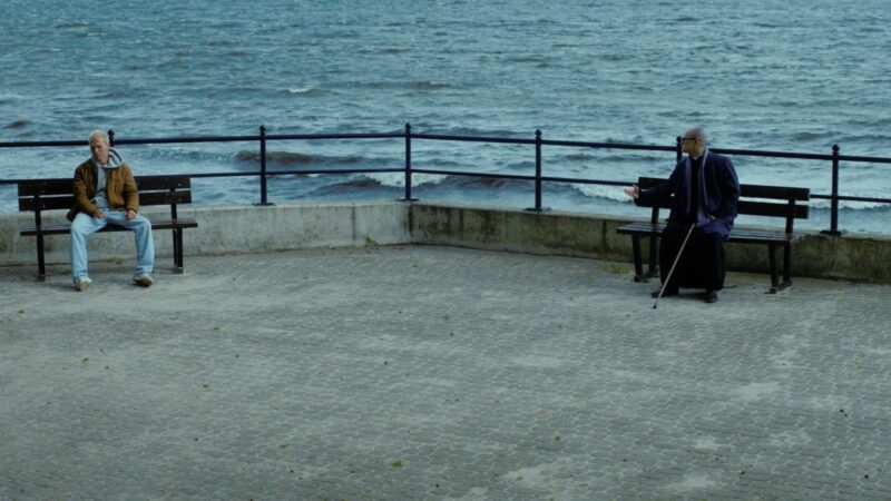 Dwójka głównych bohaterów na betonowym nabrzeżu, siędzą oddaleni od siebie na osobnych ławkach, w tle morze.