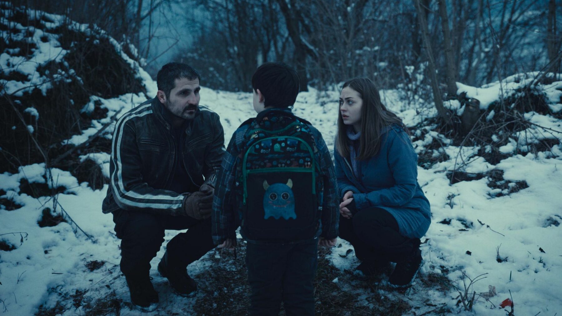 Dwoje dorosłych pochylają się nad dzieckiem z plecakiem, kadr z zimowego lasu.