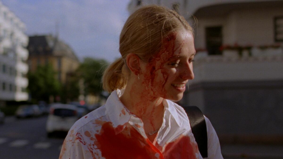 Bohaterka filmu na tle ulicy, zakrwawiona na twarzy i białej koszuli.