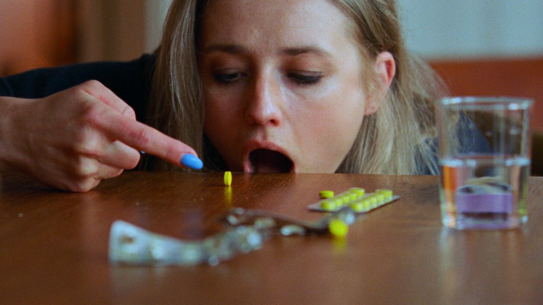 Młoda dziewczyna przed stołem z dużą ilością tabletek, łyka jedną z nich.