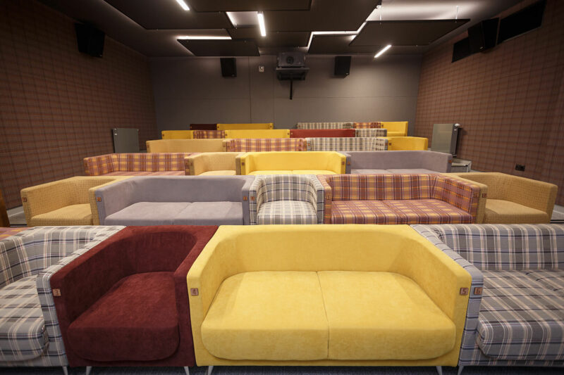 Sala kinowa na 42 fotele. Na pierwszym planie widać miejsca siedzące, na które składają się duże kolorowe fotele i kanapy. Widoczny jest też sufit z podświetlonymi elementami dekoracyjnymi o kształcie kwadratu oraz projektor znajdujący się w głębi sali.