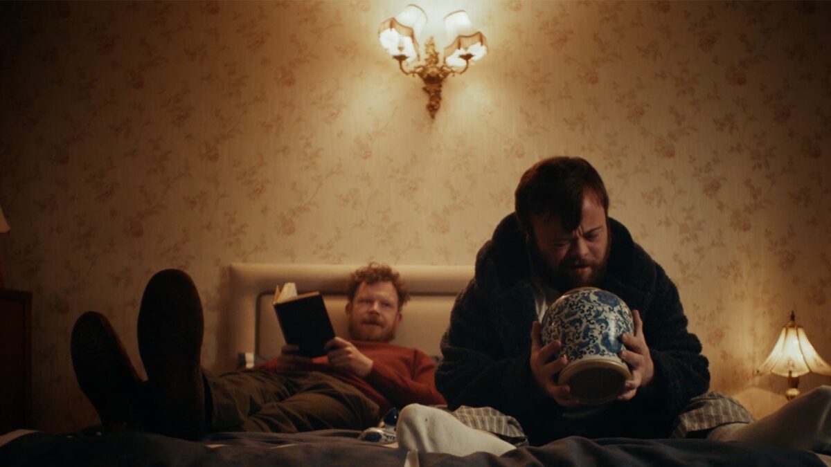 Dwóch mężczyzn na łóżku, pierwszy czyta książkę, drugi uważnie przygląda się starej wazie.