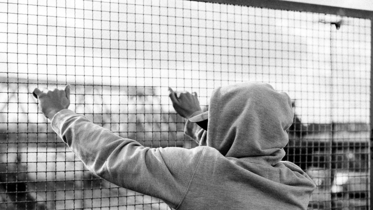 Młody chłopak stojący w kapturze tyłem, opiera się o siatkę.