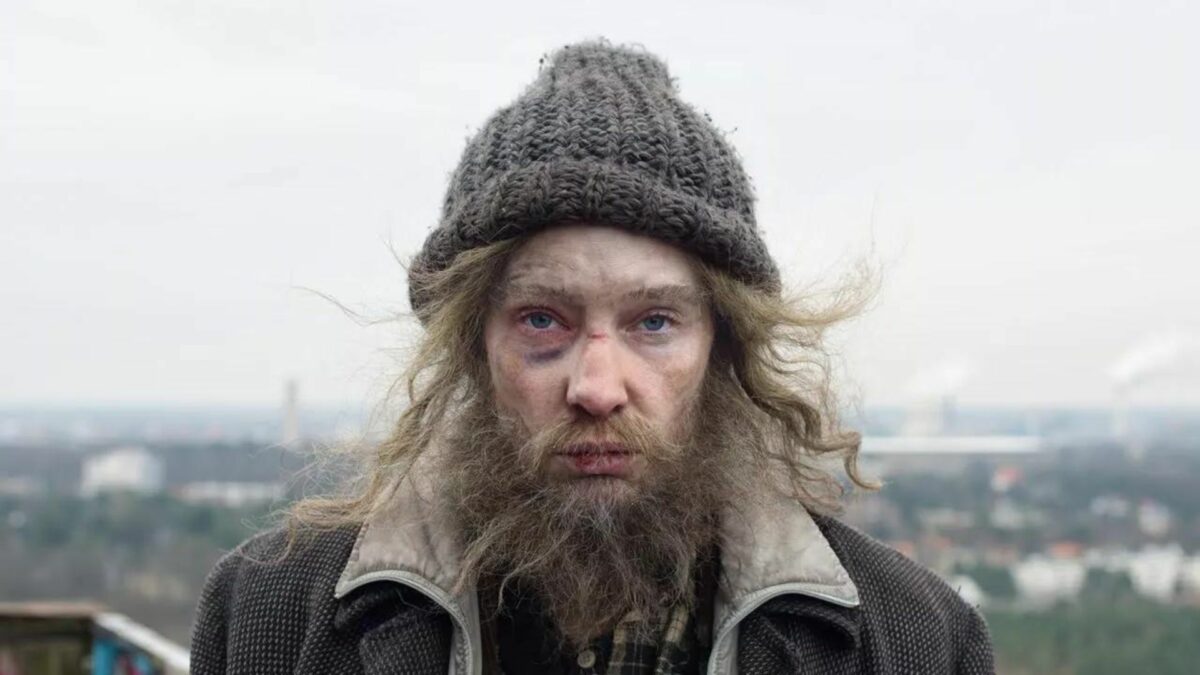 Kobieta przebrana za bezdomnego, w potarganych ciuchach oraz sztucznej brodzie.