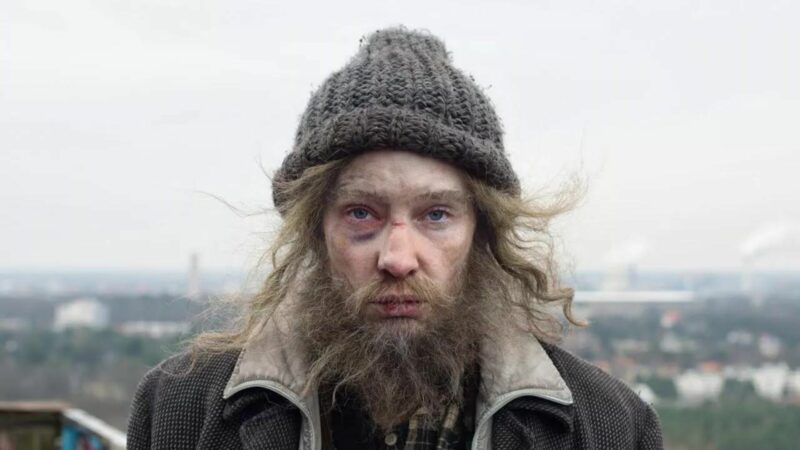 Kobieta przebrana za bezdomnego, w potarganych ciuchach oraz sztucznej brodzie.