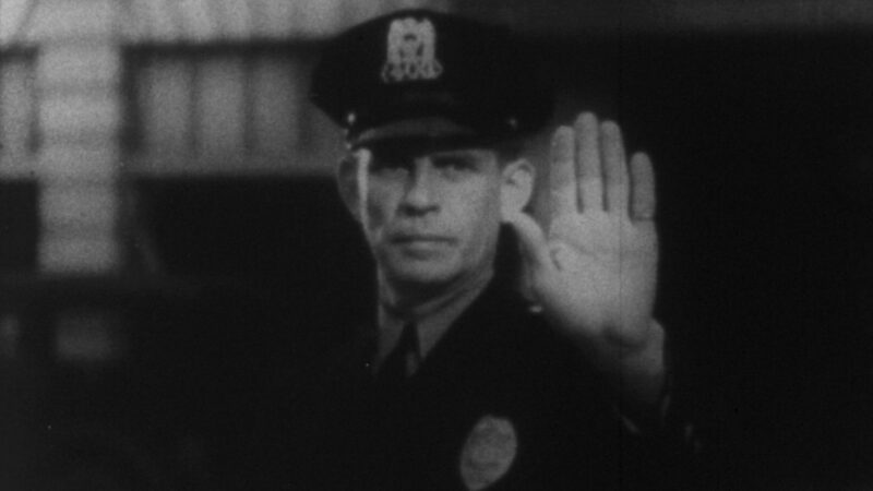 Czarnobiały kadr z policjantem z wyciągniętą dłonią na znak stop.