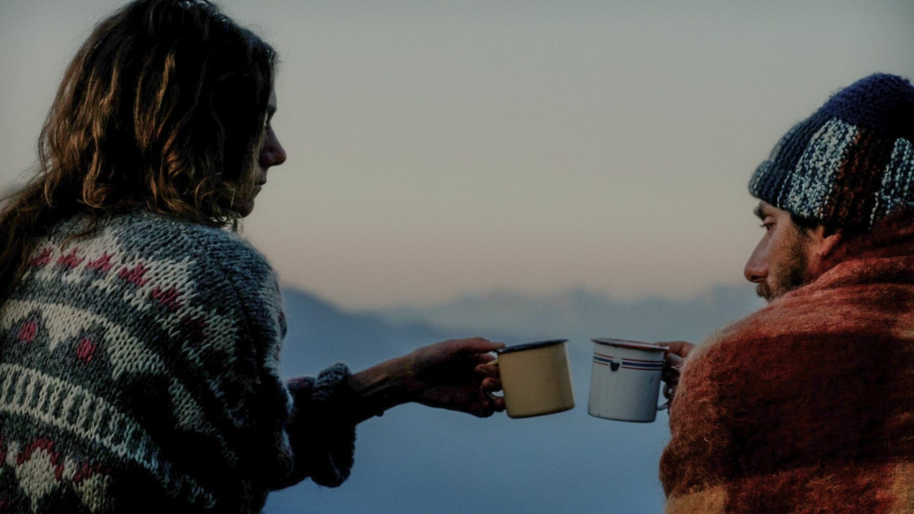 Kobieta i mężczyzna ciepło ubrani, z kocami podczas górskiego poranka, w dłoniach mają kubki z gorącym napojem.