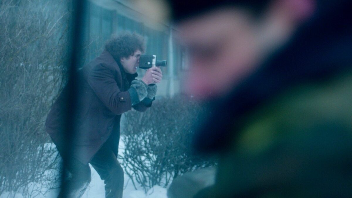 Na pierwszym planie rozmazana sylwetka mężczyzny, w tle młody chłopka z kamerą w ręku.