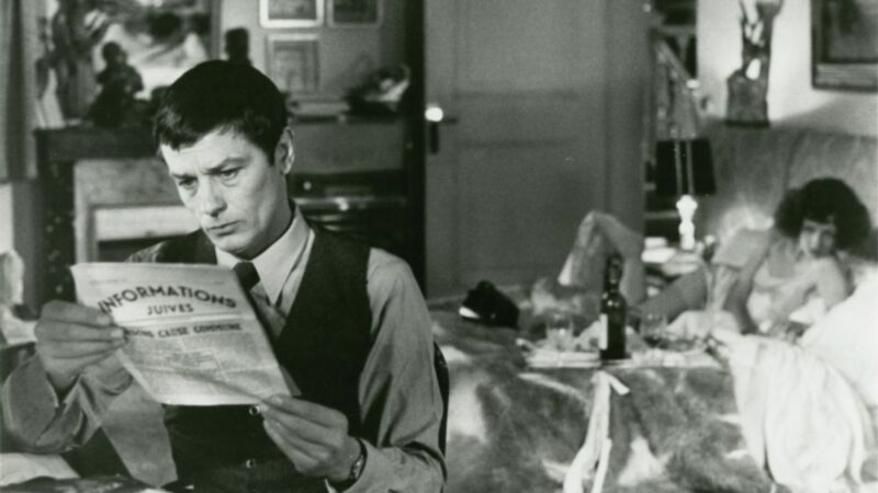 Mężczyzna czytający gazetę, na drugim planie znajduje się kobieta na łóżku, ma wino i kieliszki.