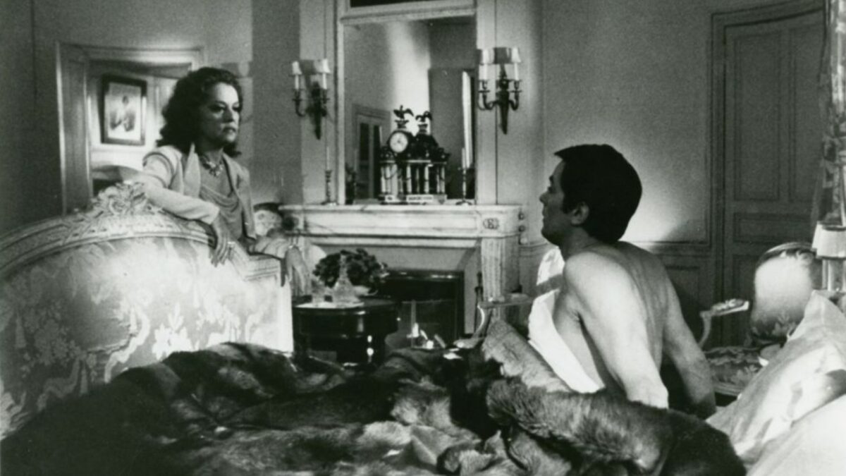 Kadr z sypialni, mężczyzna w łóżku rozmawia z kobietą opartą o ramę łóżka.