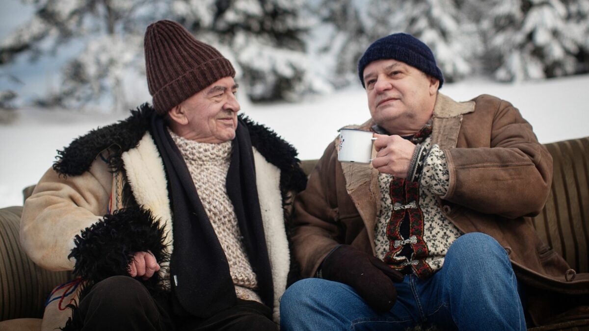 Dwóch starszych mężczyzn w zimowej scenerii, w czapkach i szalikach, jeden z nich trzyma kubek z ciepłym napojem.