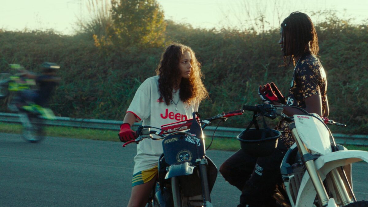 Dwoje młodych ludzi rozmawia ze sobą na zatrzymanych motorach, w tle widać innego motocyklistę jadącego na tylnim kole.