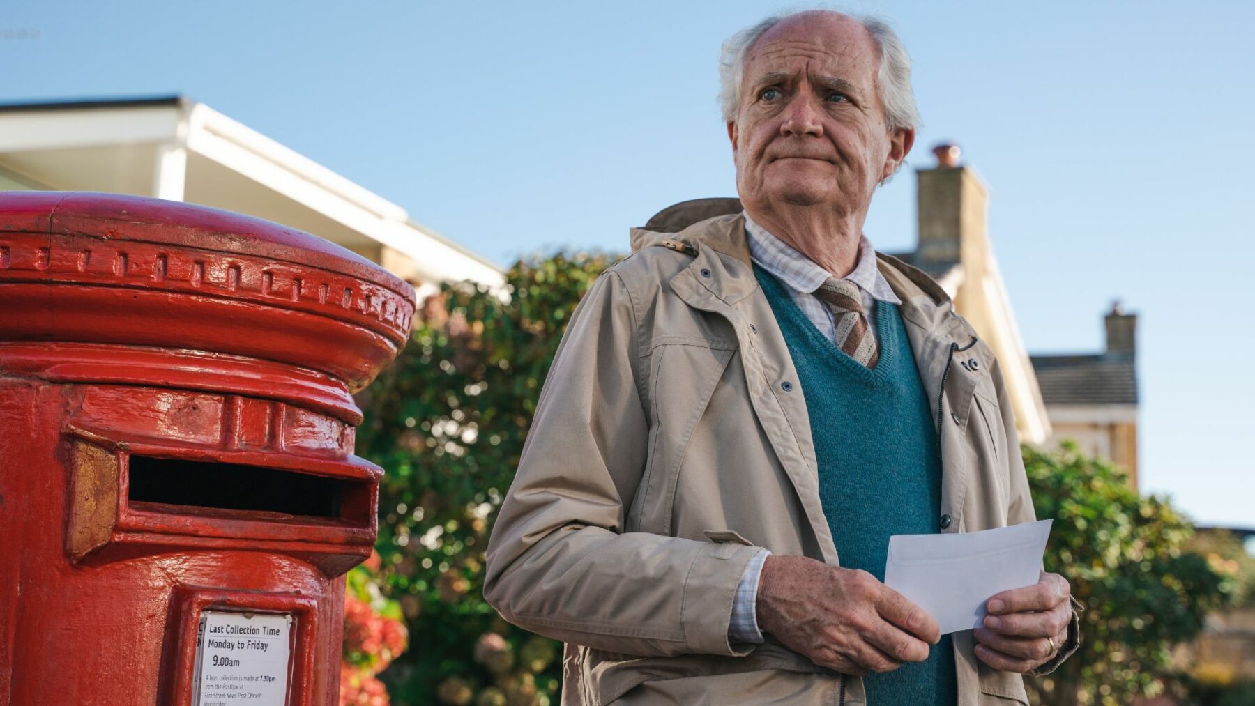 Starszy mężczyzna stojący z listem w dłoniach przed skrzynką pocztową.