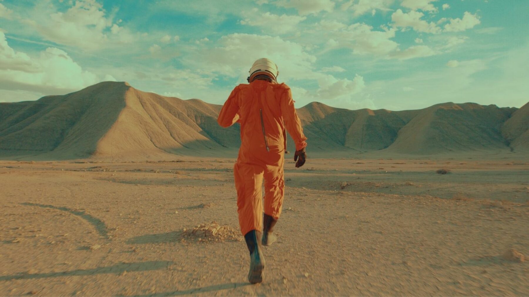 W kadrze mężczyzna biegnący po pustyni, ma na sobie pomarańczowy skafander.