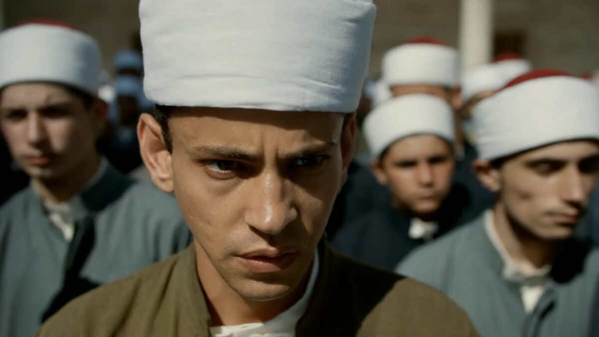 Na przednim planie młody muzułmanin, za nim grupa mężczyzn, wszyscy na głowie noszą fez.