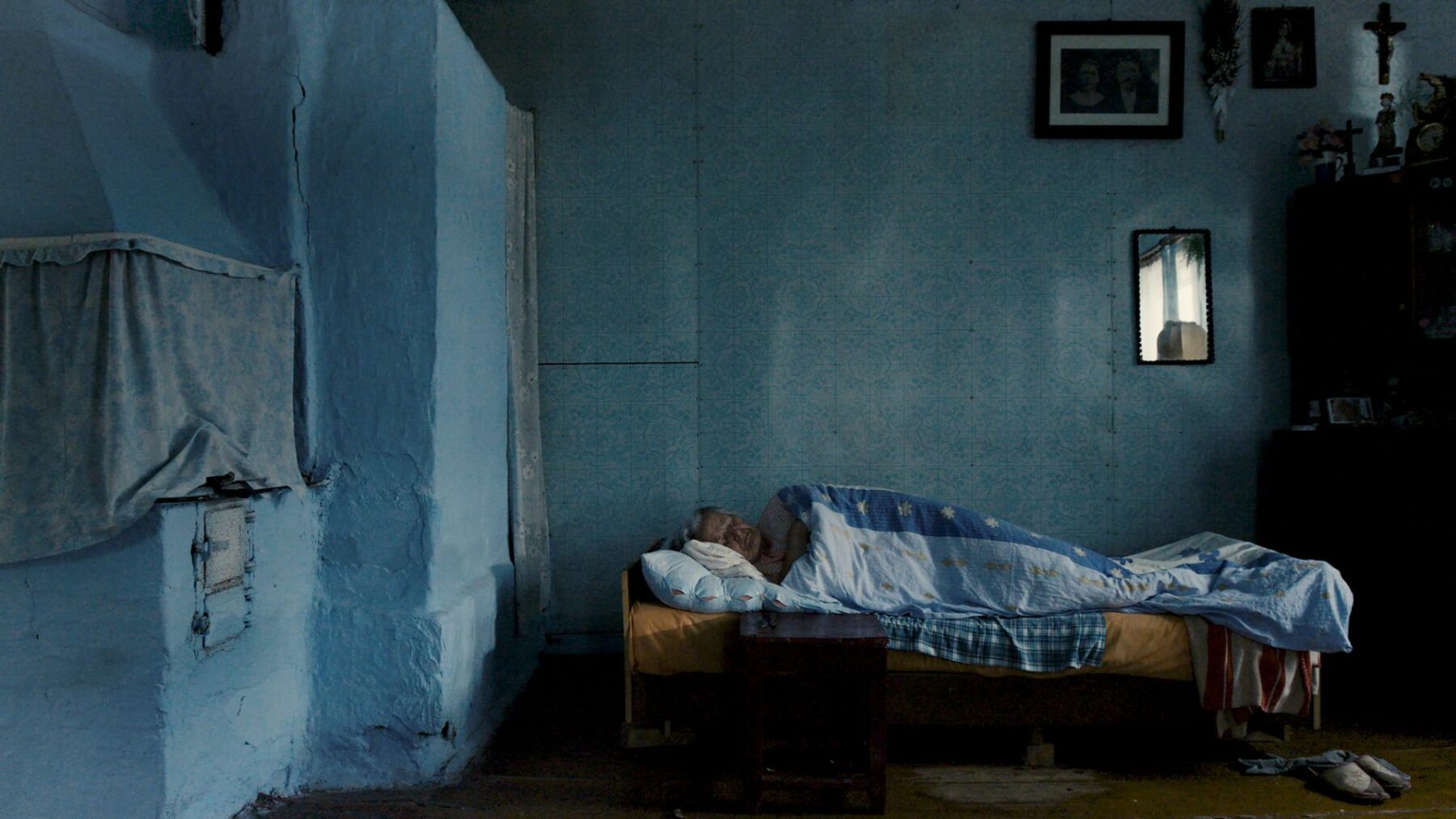 Kadr w tradycyjnym domu Polski Wschodniej, na pierwszym planie łóżko a na nim starsza kobieta.