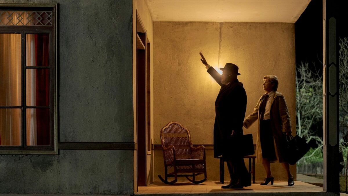 Dwie osoby stoją wieczorem na ganku. Są ubrane bardzo elegancko, w ciemne płaszcze. Na ganku świeci się mocne pomarańczowe światło. Jedna z postaci unosi rękę w geście powitania w stronę drzwi