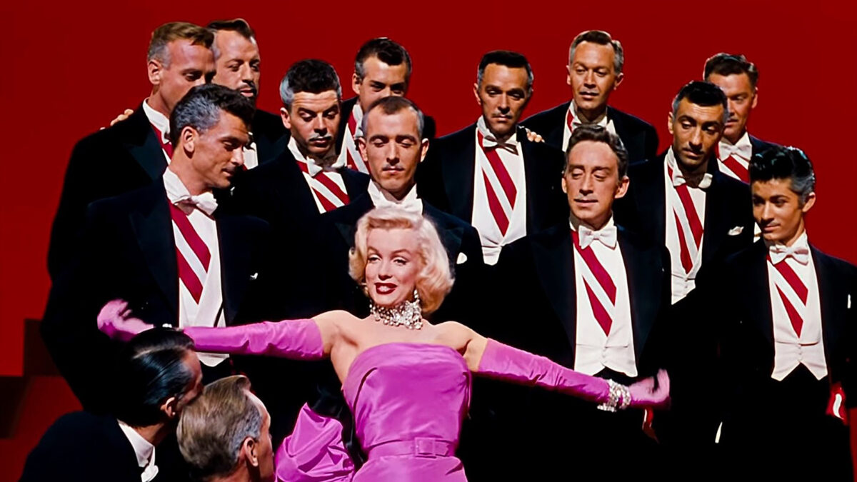 Kobieta ubrana w wieczorową różową suknię tańczy otoczona przez mężczyzn w garniturach