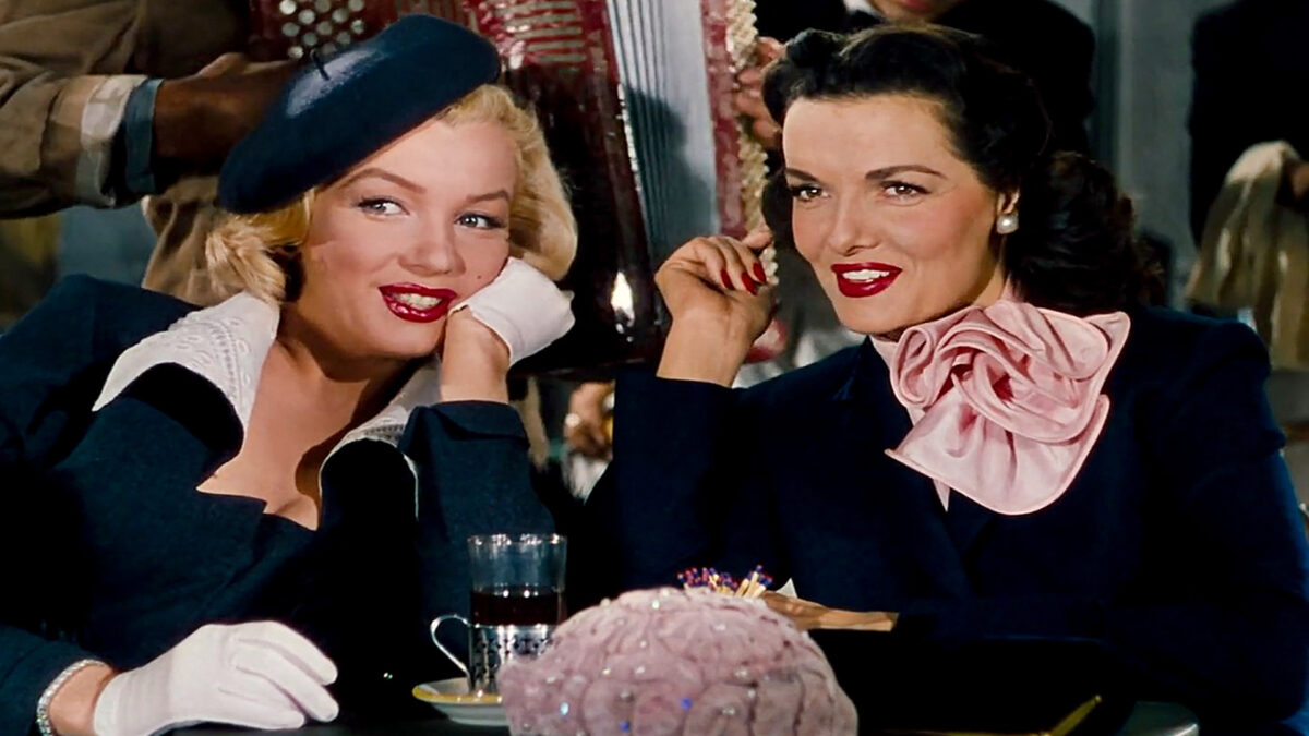 Dwie gustownie ubrane kobiety nachylają się ku sobie pogrążone w rozmowie. Obie patrzą w jednym kierunku