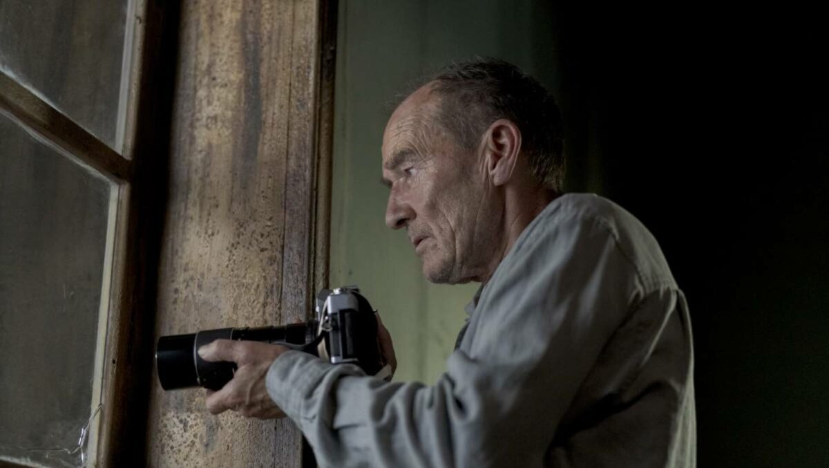 Mężczyzna z aparatem, który posiada bardzo długi obiektyw, spogląda przez okno. Pomieszczenie jest szarobure, bardzo ciemne. Mężczyzna sprawia wrażenie, jakby kogoś podglądał