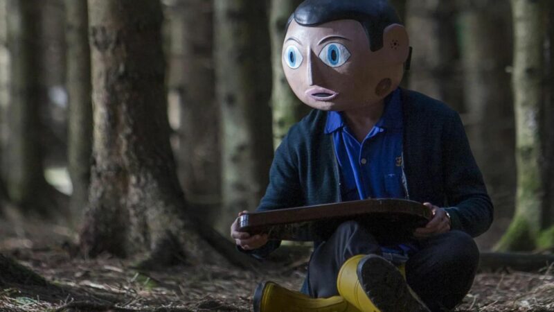 Główny bohater w masce, siedzi na ściółce w lesie.