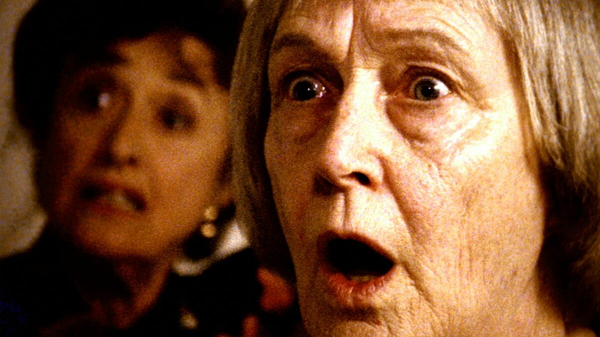 Przerażona starsza kobieta za nią w dalszym planie kolejna kobieta.