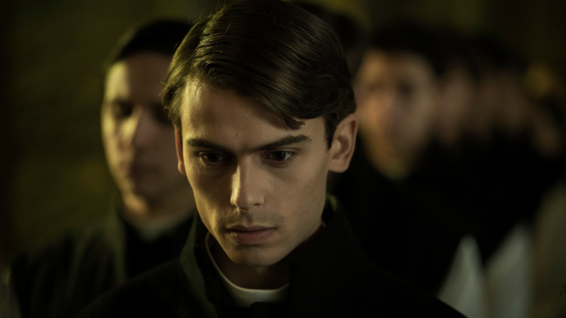 Zblizenie na twarz młodego mężczyzny, za nim widać kolejne osoby.