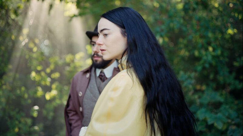 Młoda kobieta w ogrodzie a za nią widać mężczyznę.