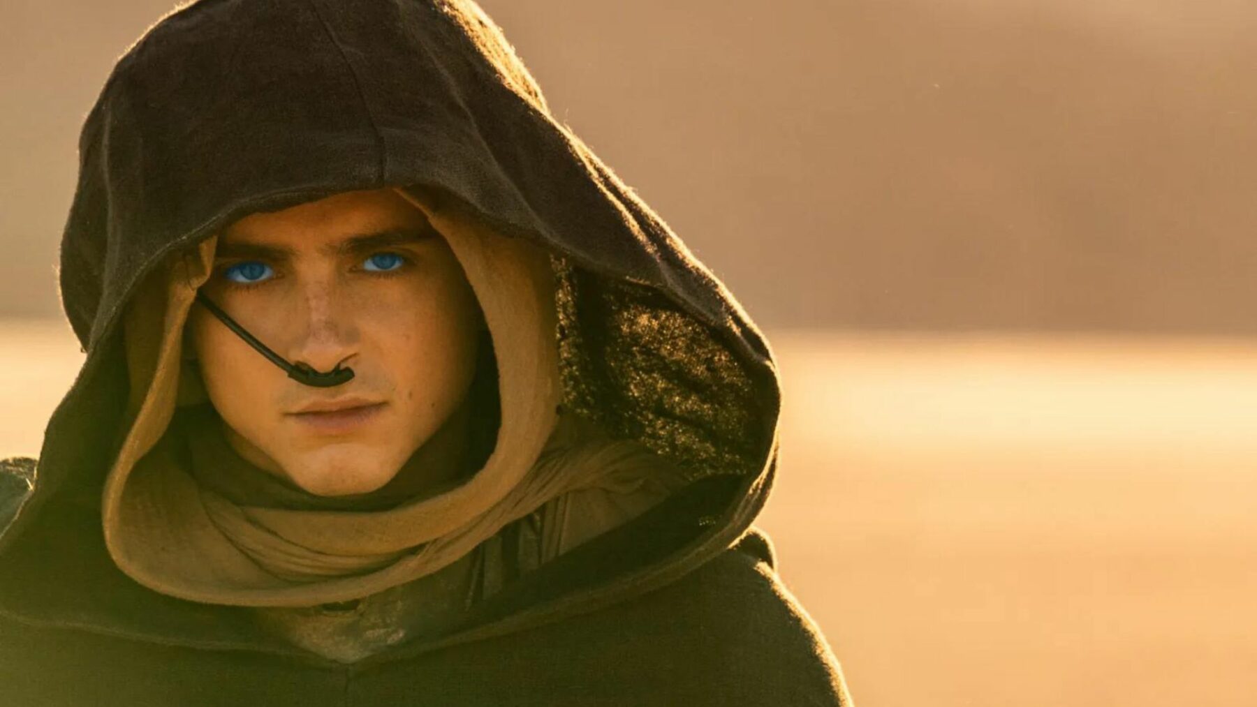 Młody chłopak na pustyni, ubrany w płaszcz z dużym kapturem.