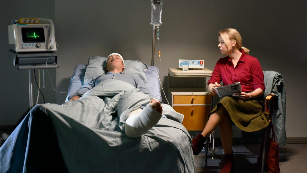 Poturbowany mężczyzna na łóżku szpitalnym a obok odwiedzająca go kobieta.