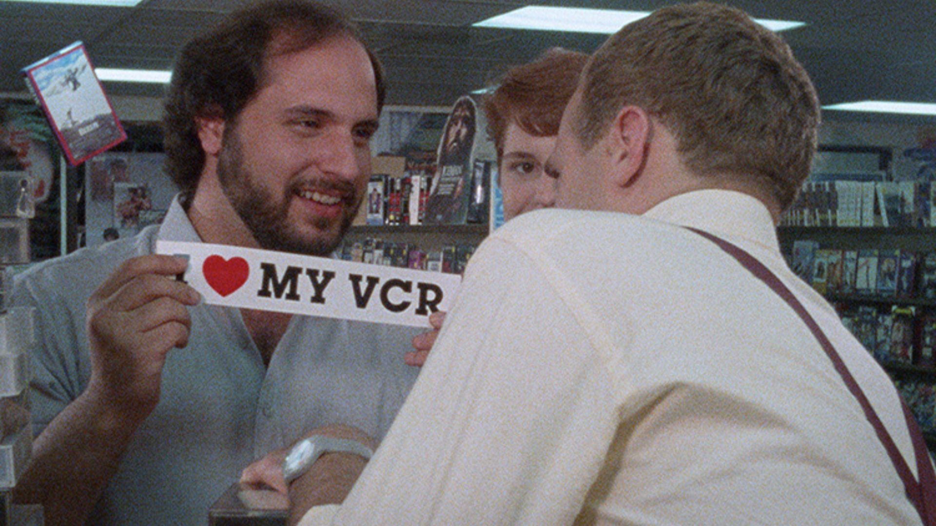 Mężczyzna pokazujący kolegą z pracy kartke z napisem I LOVE MY VCR
