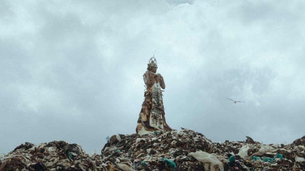 Kadr przedstawia tajemniczą osobe w kostiumie na wysypisku śmieci.