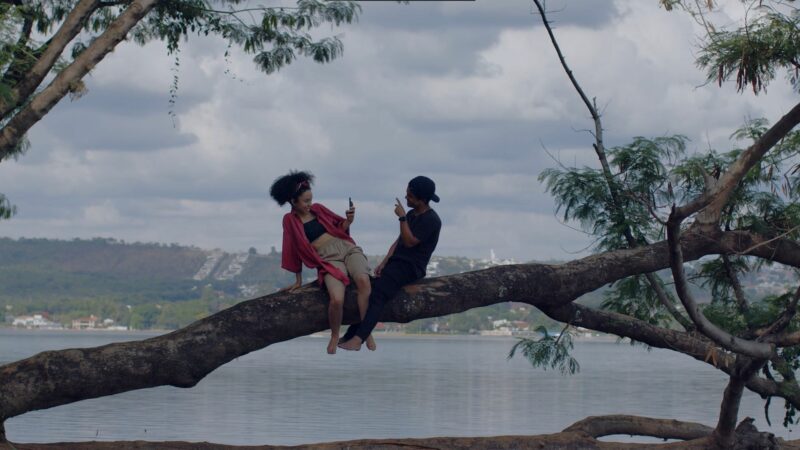 Kadr przedstawia dwie młode osoby spędzające czas nad rzeką, siedzą na gałęzi drzewa.