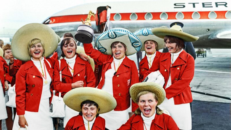 Grupa kobiet na lotnisku ma na sobie czerwono-białe ubrania i meksykańskie kapelusze, z czego jedna z nich wnosi w górę trofeum