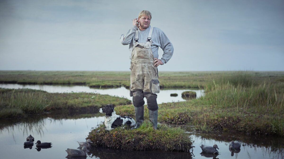 Mężczyzna i pies na malutkiej wysepce otoczonej kaczkami, pływającymi na powierzchni wody