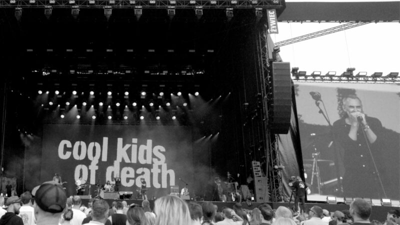 Czarno-biały kadr ze sceną koncertową z napisem "Cool kids of death" oraz zebraną publicznością