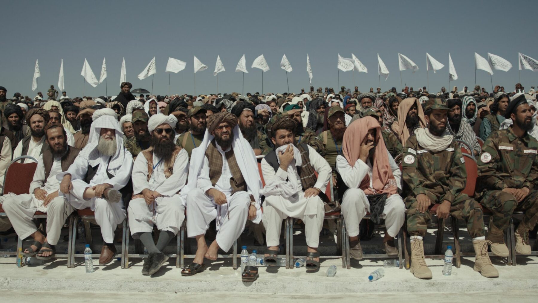 Duża grupa mężczyzn siedzących na krzesłach w kilku rzędach, a nad nimi powiewają białe flagi