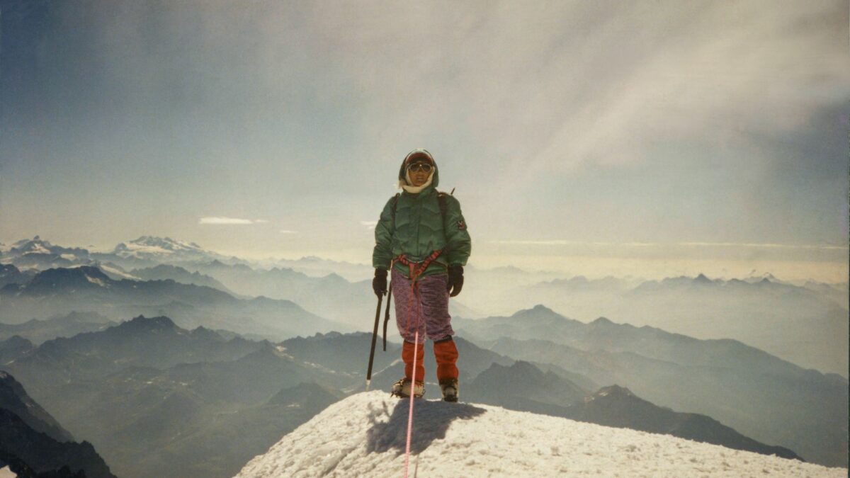 Kobieta na szczycie góry, za nią szeroki widok na pasmo górskie.