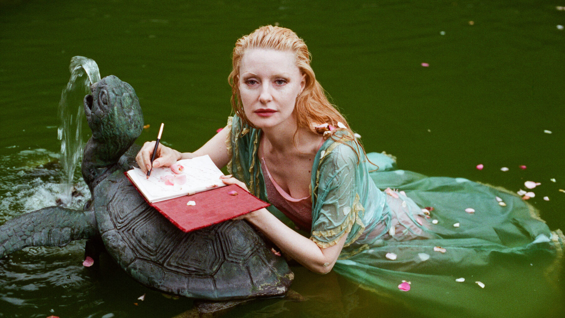 Kobieta w wodzie pisze w zeszycie opierając się o fontannę w kształcie żółwia