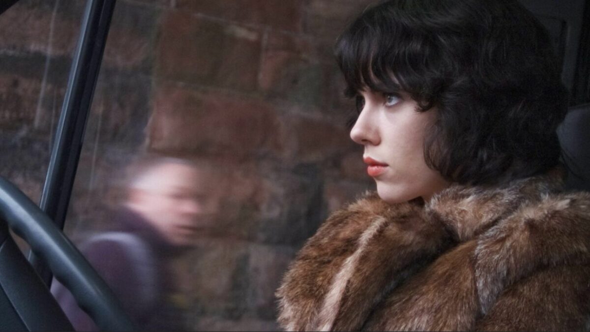 Kobieta prowadząca samochód, za szybą widać sylwetkę mężczyzny.