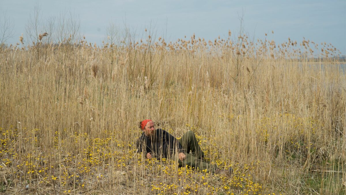 Kadr przedstawia mężczyznę siedzącego na ziemi wśród nadrzecznej roślinności i kwiatów