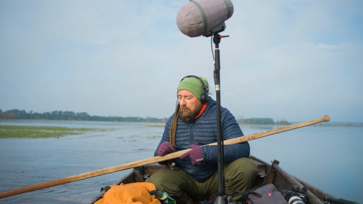 Mężczyzna w słuchawkach siedzi w łodzi i rejestruje dźwięki otoczenia za pomocą dużego mikrofonu