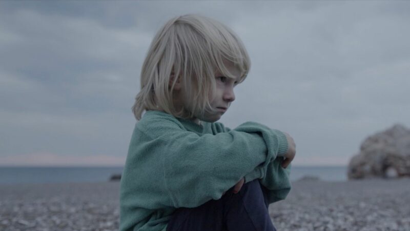 Chłopiec w dużej bluzie na kamiennej plaży.