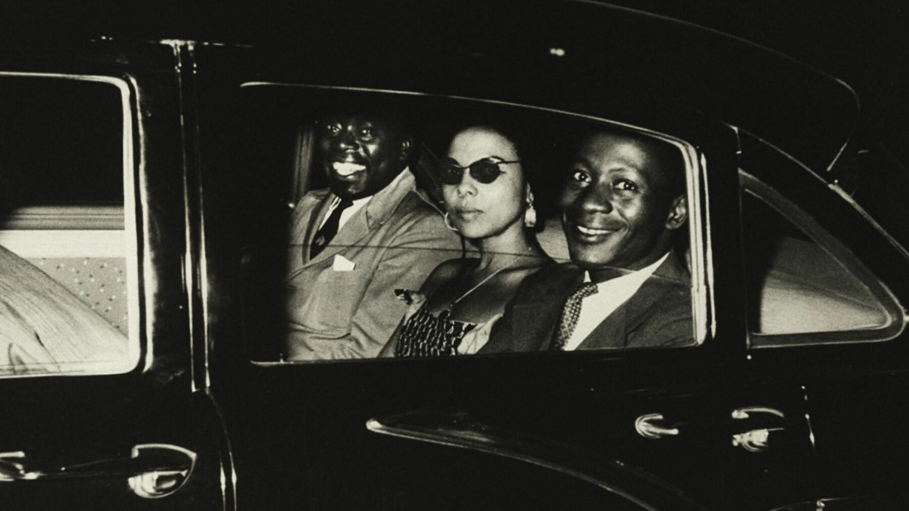 Czarno-biały kadr przedstawiający dwóch mężczyzn oraz kobietę na tylnych siedzeniach w samochodzie