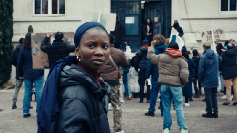 Zbliżenie na młodą kobietę, za jej placami odbywa się prostest na ulicy.